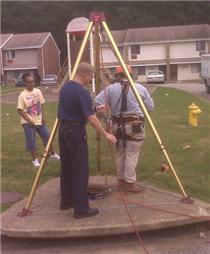 Chattanooga firefighters rig up a tripod  and attach a harness to Capt. Tallent before lowering him down into the drain.  Ms. Wallace (in yellow T-shirt) stands nearby to watch the rescue operation.